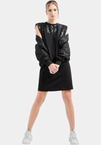 Abito nero con spalle scoperte, caratterizzato da un pannello lucido a fantasia sul petto e maniche voluminose e strutturate. Abbinato a sneakers bianche.