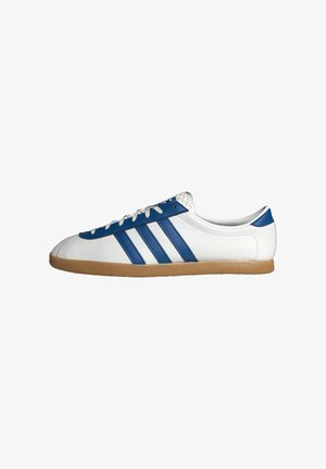 Zapatilla baja blanca con rayas azules, acentos azules, cordones blancos y una suela de goma marrón claro, mostrada de perfil sobre un fondo blanco.