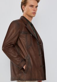 Chaqueta de cuero marrón con un diseño entallado, que presenta un cuello con muesca, botones frontales, un bolsillo con cremallera y una textura suave.