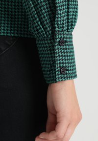 Grön och svart houndstooth-mönstrad skjortärm med två svarta knappar, med en slät tygtextur och en skräddarsydd passform.