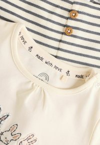 Two garments: a cream top with bunny graphics and a ribbed striped top in cream and navy, featuring wooden buttons and a logo tag.
