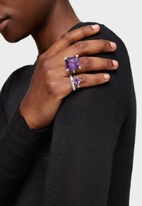 Anillos de ametista con una gran piedra rectangular y una más pequeña redonda en un engaste de plata, llevados en una mano contra una blusa negra.