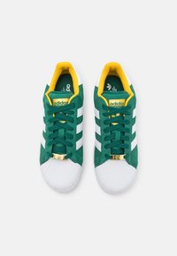 Green suede sneakers with white stripes and toe cap. Yellow lining and gold-accented branding on the tongue and lace area.