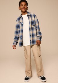 Camicia a quadri blu e crema sopra una t-shirt bianca, pantaloni cargo color sabbia e sneakers nere. L'outfit presenta una vestibilità comoda e tasche.