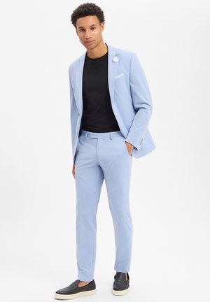 Mann in hellblauem Anzug mit schwarzem Hemd und schwarzen Slippern, der mit einer Hand in der Tasche vor weißem Hintergrund steht.