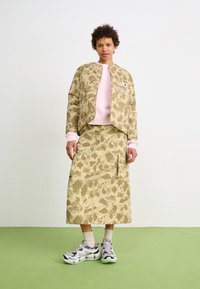 Giacca bomber con motivo mimetico e gonna coordinata in tonalità beige. Il modello indossa una felpa rosa chiaro sotto con tasche cargo e sneaker.