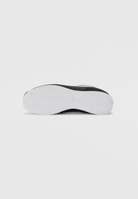 Fekete sneaker texturált felsőrésszel, fehér cikk-cakk talppal és minimalista formatervezéssel. A talp alsó részletei és logója látható.