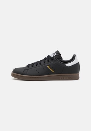 Zwarte Adidas Stan Smith sneaker met bruine zool, witte hielkap, zwarte veters en gouden "Stan Smith" tekst aan de zijkant.