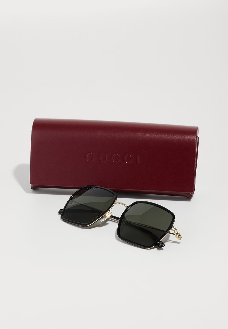 Occhiali da sole neri quadrati con lenti scure, dettagli dorati, affiancati da un astuccio in pelle bordeaux stampato con "GUCCI." Texture liscia.