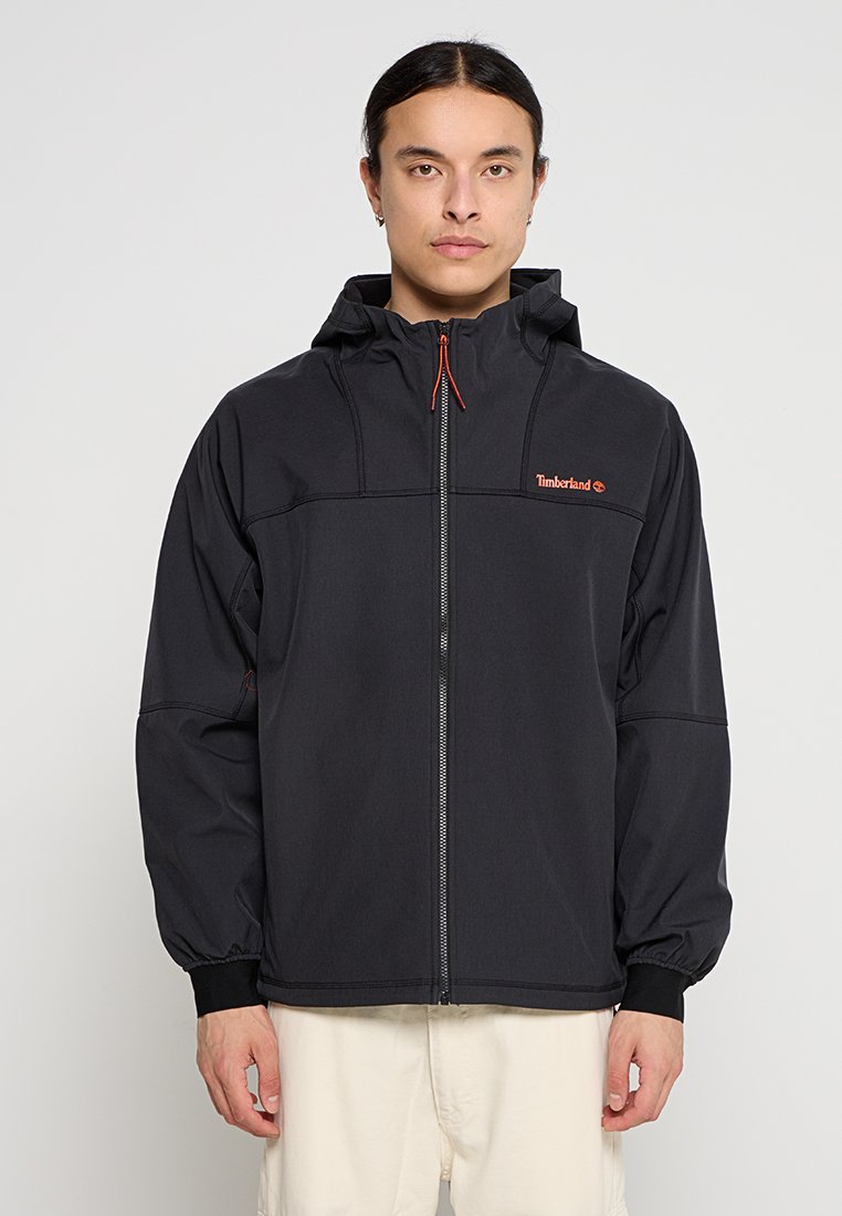 Chaqueta ligera negra con cremallera frontal, capucha ajustable y puños elásticos. Presenta un pequeño logotipo rojo de Timberland en el pecho.