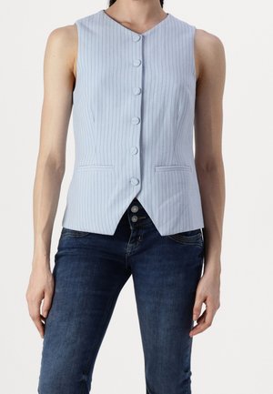 Kobieta ubrana w jasnoniebieską kamizelkę bez rękawów w cienkie prążki z guzikami, zestawioną z ciemnoniebieskimi jeansami na jednolitym tle.
