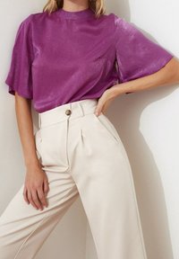 Top corto a maniche corte in satin viola con collare alto e taschino sul petto, abbinato a pantaloni beige a vita alta con un unico bottone e pieghe.