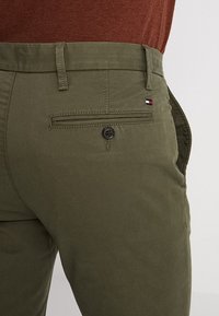 Pantaloni verde oliva con frontale piatto, tasca posteriore con bottone e piccolo'etichetta logo vicino alla tasca. Realizzati in tessuto di cotone.