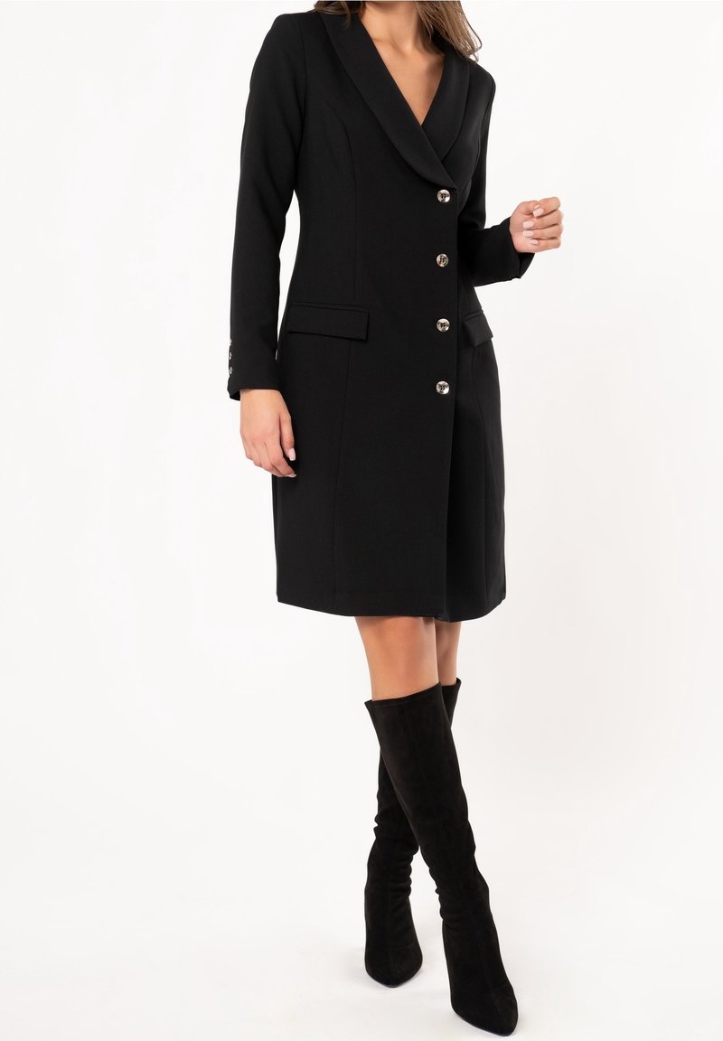 Donna che indossa un abito blazer nero al ginocchio con bottoni argentati e stivali in camoscio neri alti fino al ginocchio, su uno sfondo bianco.