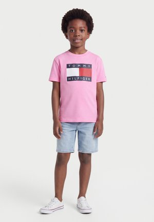 Garçon debout et souriant, portant un t-shirt rose Tommy Hilfiger, un short en jean bleu clair et des baskets blanches sur fond blanc.