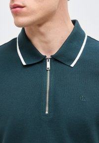 Κοντινό πλάνο ενός ατόμου που φοράει σκούρο πράσινο πουκάμισο με γιακά και λευκή επένδυση, με ασημένιο φερμουάρ εμπρός, που δείχνει μερικώς το λογότυπο cK.