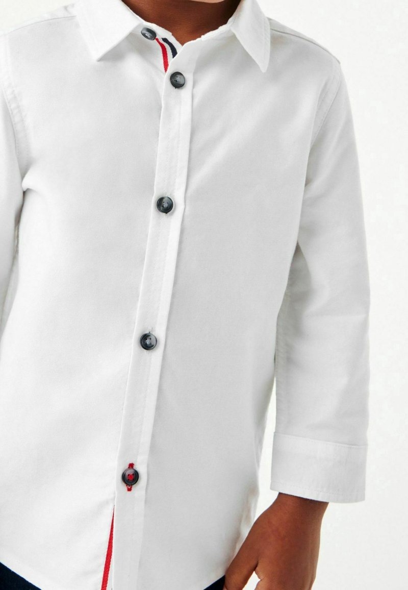 Enfant portant une chemise blanche à manches longues avec des boutons noirs et des détails rouges à l’intérieur de la patte de boutonnage.