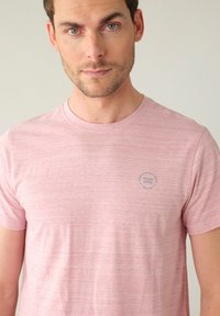 Helles rosa Baumwoll-T-Shirt mit Rundhalsausschnitt, kurzen Ärmeln und einer dezenten Textur. Mit einem blauen kreisförmigen Logo auf der linken Brust.