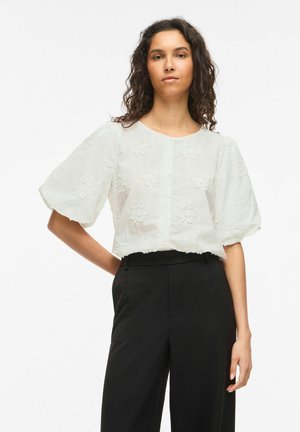 Frau mit lockigem Haar trägt eine weiße, mit Blumen bestickte Bluse mit Puffärmeln und schwarze hoch taillierte Hose, steht mit einer Hand in der Hüfte.
