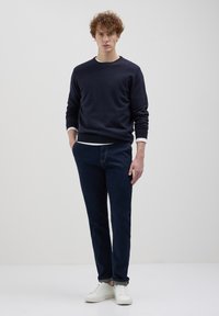 Marineblauer Pullover mit Rundhalsausschnitt, weißes Unterhemd an den Ärmelbündchen sichtbar, kombiniert mit dunkelblauen Jeans und weißen Sneakers. Einfaches Design, lässige Passform.