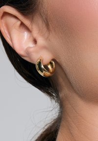 Złota kolczyk w kształcie koła, o wypolerowanej powierzchni, z lekko przekręconym detalem, spoczywający na płatku ucha, eksponujący blask i gładką teksturę.