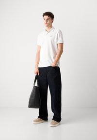 Hvid bomulds polo skjorte med korte ærmer og lille rødt logo, parret med mørkeblå brede bukser og beige sneakers, mens der holdes en sort taske.