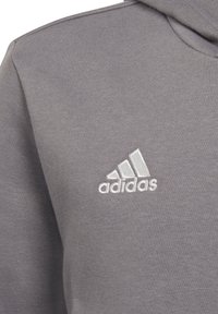 Felpa grigia con cappuccio realizzata in tessuto testurizzato. Presenta un logo Adidas ricamato in bianco sul petto. Design semplice con cappuccio.