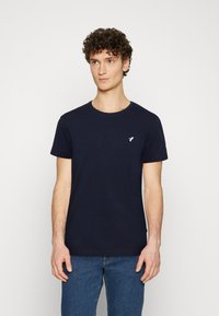 Navy blue cotton t-shirt with short sleeves and a crew neckline, featuring a small white bird logo on the chest. Worn with blue jeans.
