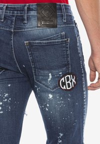 Jean en denim bleu avec une pièce en cuir, des détails déchirés et un logo "CBX" brodé sur la poche arrière.