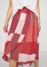 Jupe à motifs rouges et roses avec un design en couches, présentant diverses textures et un ourlet asymétrique. Tissu léger, taille élastique.