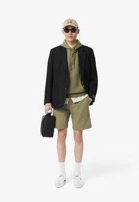 Blazer negro sobre una sudadera verde y una camisa de rayas, combinado con un pantalón corto a juego. Los accesorios incluyen una gorra beige, gafas de sol y un bolso negro acolchado.