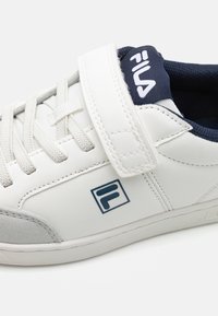 Bijela tenisica s mornarsko plavim detaljima, teksturirana koža, vezice i remen na čičak. Ima sivi ante detalj na vrhu prstiju i logo sa strane.