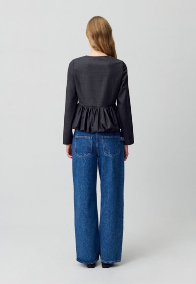 Top peplum grigio scuro con maniche lunghe e vita arricciata; abbinato a jeans in denim blu larghi con tasche posteriori e taglio dritto.