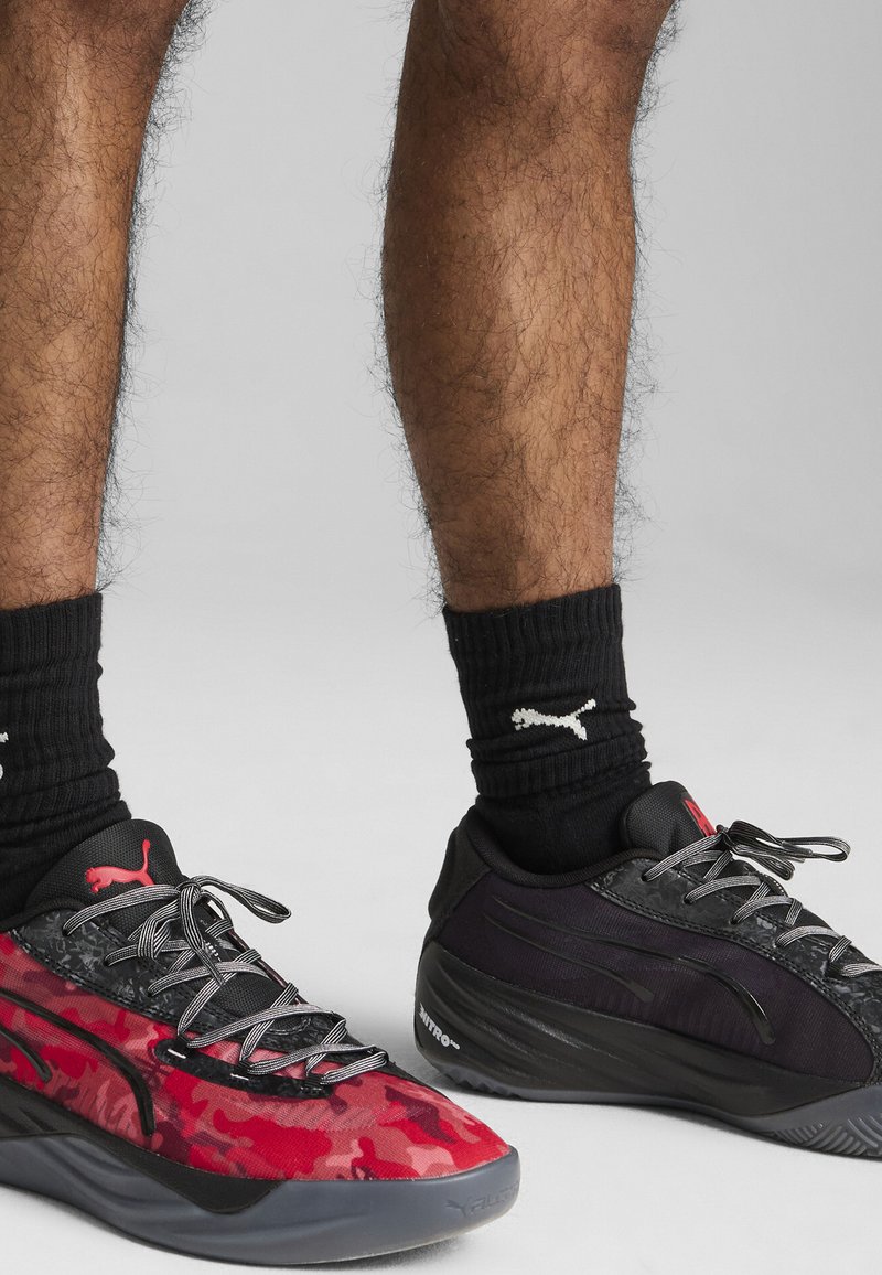 Baskets de sport en camouflage noir et rouge avec une maille texturée et des superpositions synthétiques, accompagnées de chaussettes noires côtelées arborant un logo blanc.