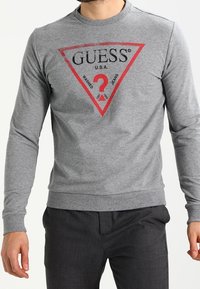 Sudadera gris de manga larga con un gráfico triangular que presenta el texto "GUESS U.S.A." en negro y rojo. Puños y dobladillo acanalados. Tejido suave.