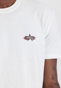 Hvid bomulds t-shirt med et lille logo, der viser et abstrakt sort stjernes design og røde penselstræk på venstre brystområde.
