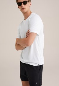 Weißes T-Shirt mit Rundhalsausschnitt und kurzen Ärmeln, kombiniert mit schwarzen Shorts, die am Saum ein kleines Logo aufweisen. Das Model trägt Sonnenbrillen.