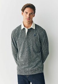 Grå hørhjerte-mønstret langærmet polo med hvid krave og blåt logo. Stoffet fremstår tekstureret. Båret over blå jeans.