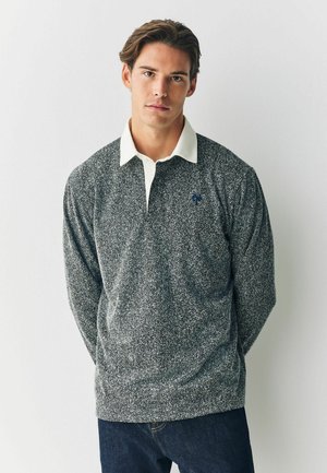 Szara longsleeve polo w wzór jodełki z białym kołnierzykiem i niebieskim logo. Materiał wydaje się być teksturowany. Nosi się do niebieskich dżinsów.
