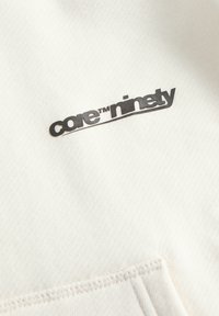 Zwart "core™ ninety" logo gedrukt op gebroken witte textuurstof boven een gestikte naad.