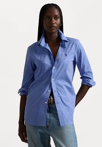 Chemise bleu clair à boutons en coton, avec un col pointu, des manches longues retroussées et un petit logo sur la poitrine gauche. Portez-la avec un jean.