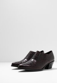 Mörkbruna klackade ankelskor med en strukturerad, krokodilliknande yta, placerade på en vit bakgrund.