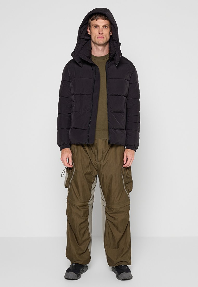 Giubbotto puffer nero con cappuccio, polsini elasticizzati e chiusura con zip, indossato sopra una maglia verde oliva e pantaloni cargo larghi verde oliva con tasche laterali.