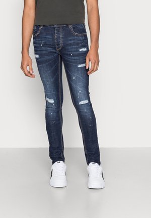 Homme portant un jean skinny bleu foncé usé avec des éclaboussures de peinture blanche et des baskets blanches Puma, debout sur un sol gris clair.