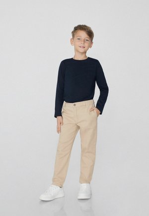 Chemise à manches longues bleu marine, pantalon beige et baskets blanches. La chemise a un col rond ; le pantalon présente des détails de bouton et de poches.