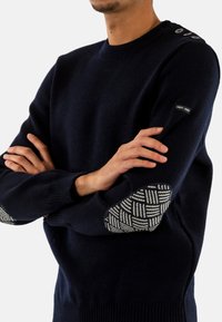 Maglione blu navy con scollatura rotonda, polsini a costine e una toppa per il gomito bianca a motivo testurizzato. Presenta bottoni decorativi sulla spalla.