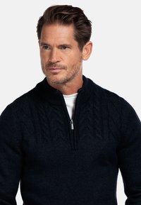 Volwassen man met kort bruin haar en stoppelbaard, gekleed in een donkerblauw kabelgebreid half-rits trui over een wit shirt, die iets naar rechts kijkt.