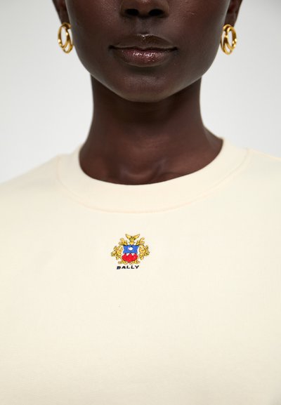 Cremefarvet sweatshirt med ribbet halsudskæring, prydet med farverigt broderet logo med et våbenskjold design og teksten "BALLY" nedenfor.