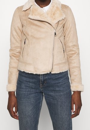 Kvinde iført beige kunstigt lammeskindsjakke med asymmetrisk lynlås, lynlåslommer i siden og blå denimjeans, stående mod en hvid baggrund.