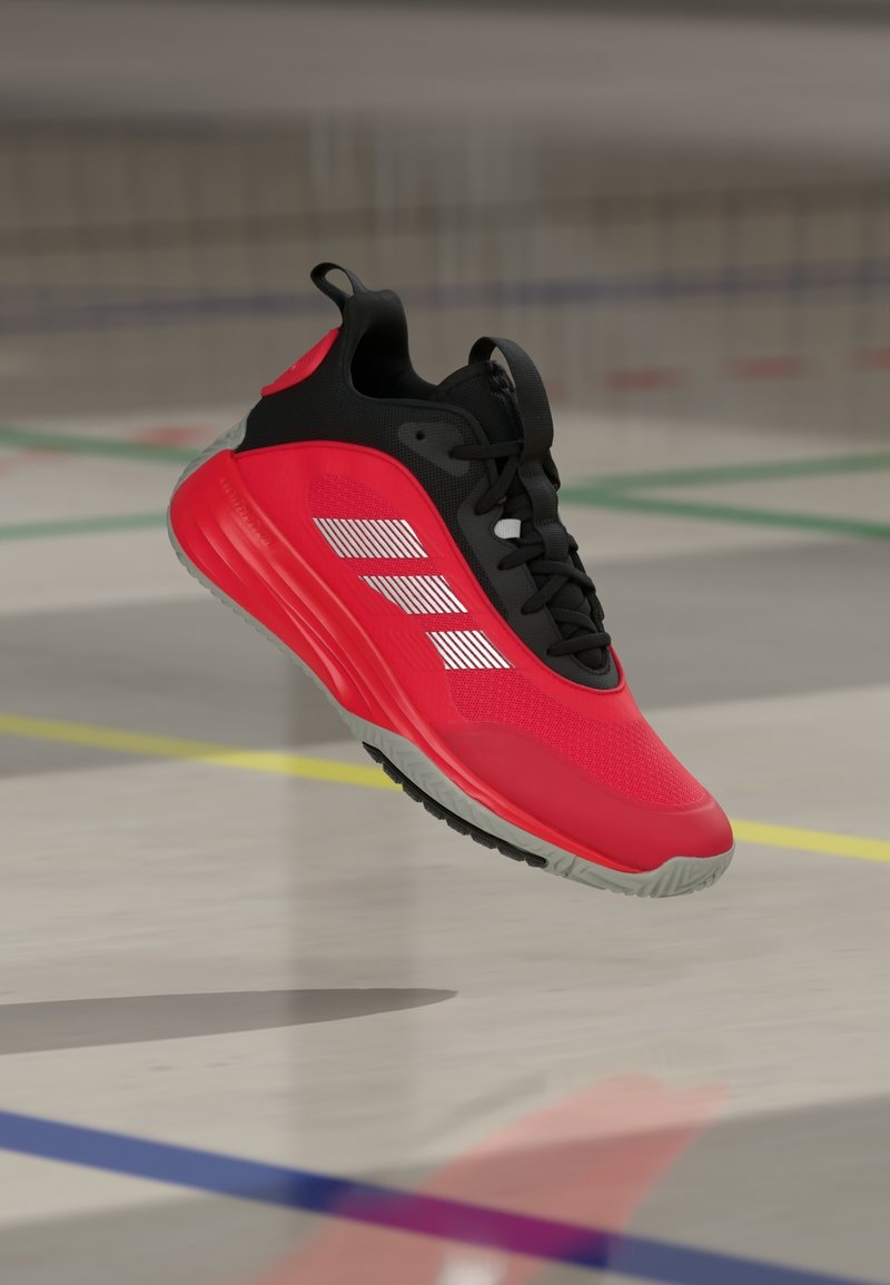 Chaussure de sport rouge et noire avec des bandes blanches suspendue au-dessus d'un sol de gym marqué de lignes colorées.